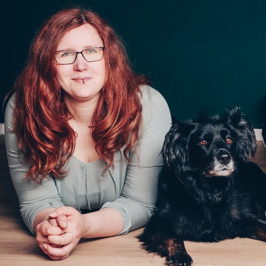 Frau mit langen roten Haaren und Brille, liegt neben einem schwarzen Hund.